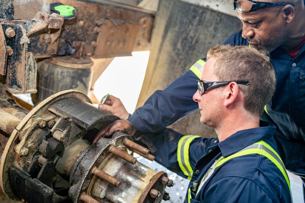 Heavy equipment mechanic Two workers in safety gear inspect a rusted industrial machine, one holding a tool near exposed bolts and a worn belt.